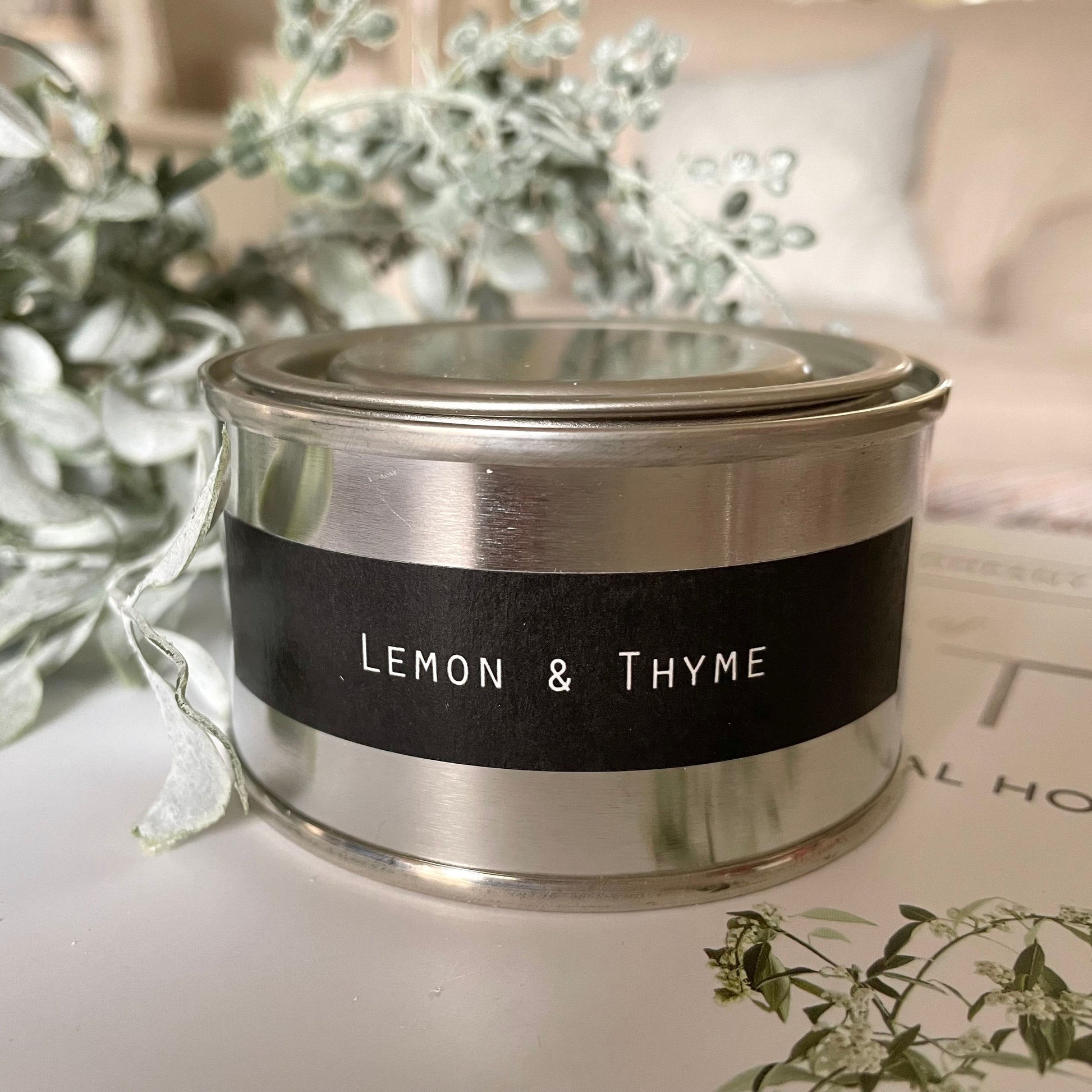 Candle in a metal container with 'Lemon & Thyme' label on a surface with greenery.