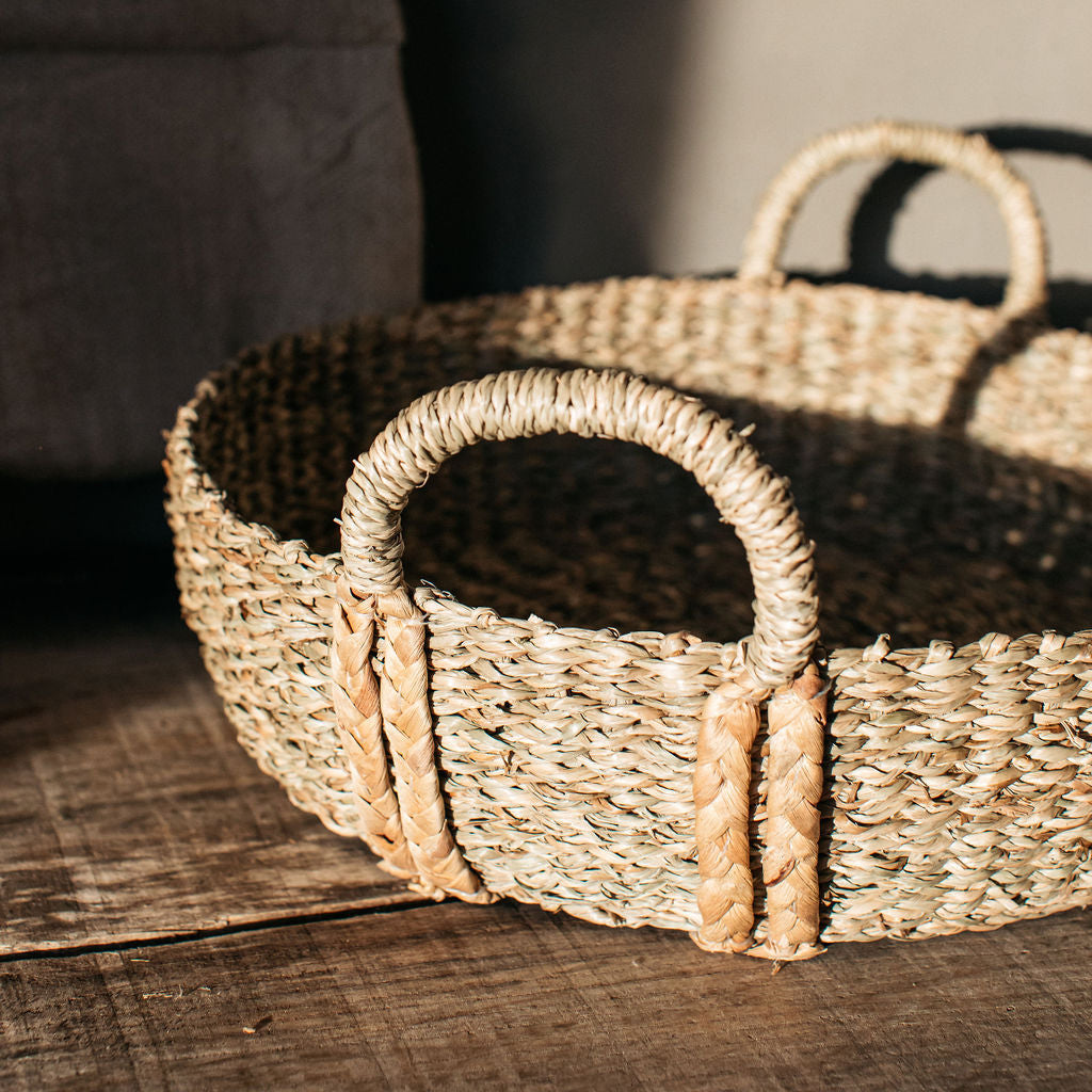 Handwoven natural seagrass basket with plaited handles, styled on a coffee table with books and candles for a warm, rustic look.