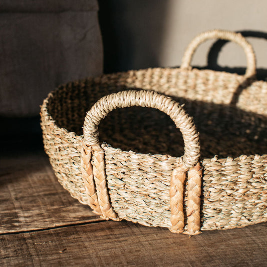 Handwoven natural seagrass basket with plaited handles, styled on a coffee table with books and candles for a warm, rustic look.