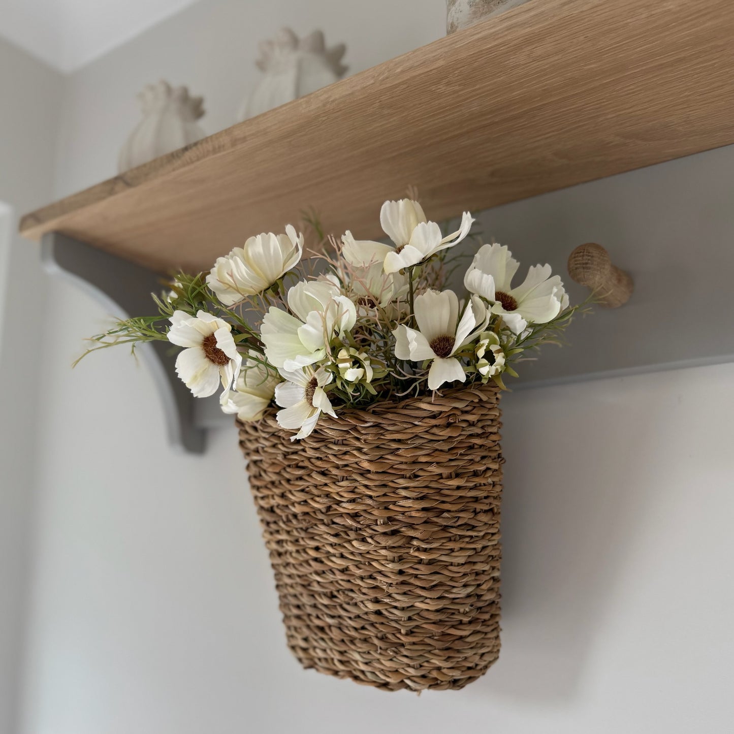 Woven basket with white flowers on a wooden shelf against a light wall.