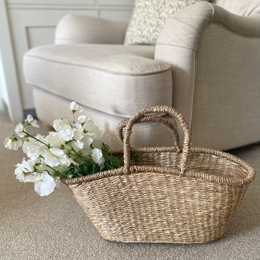 Handwoven seagrass shopper basket with natural texture styled with white sweet peas. On a beige textured carpet.
