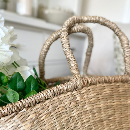 Handwoven seagrass shopper basket with natural texture. Styled on a cream sofa with a bunch of white sweet pea stems.