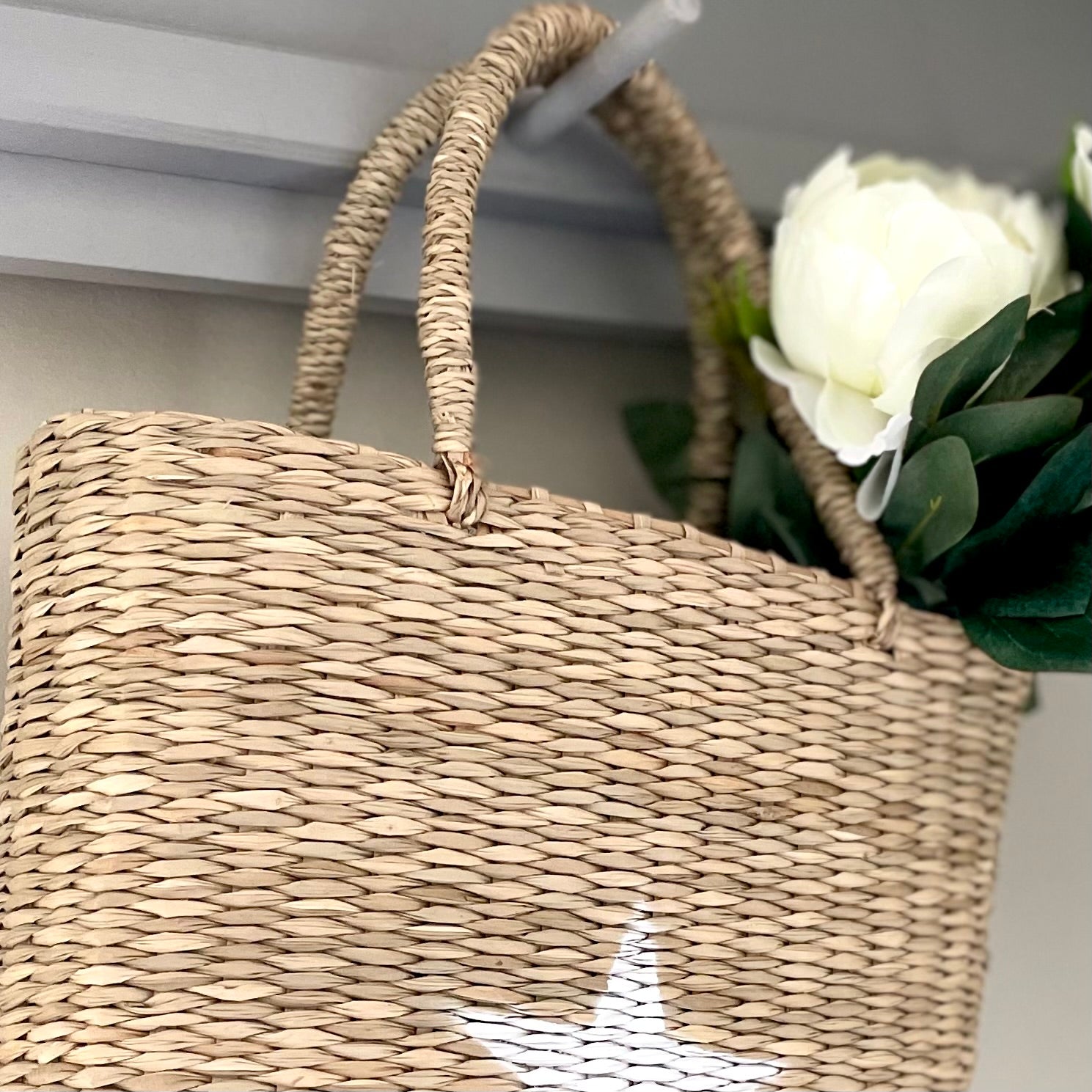Hand-painted white star seagrass tote bag with natural woven handles, styled with faux flowers hanging on a peg shelf.