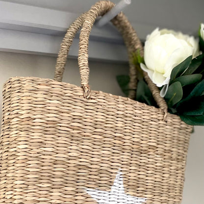 Hand-painted white star seagrass tote bag with natural woven handles, styled with faux flowers hanging on a peg shelf.
