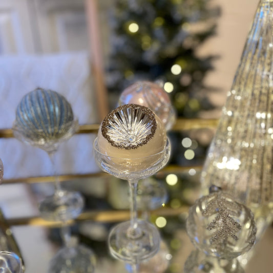Set of three pearlescent Christmas baubles with delicate cut out detailing. Styled on a glass table with a Christmas tree with pretty lights.