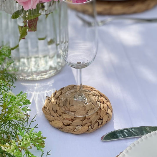 Set of 4 round woven hyacinth coasters arranged on a tabletop, showing there natural texture and warm, organic colour. Styled on a garden dining table with a vase of roses.