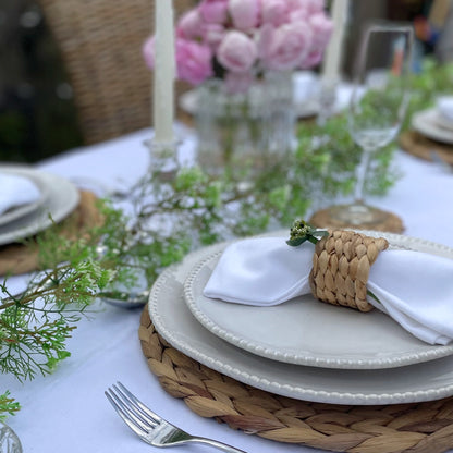 Round woven water hyacinth placemat showing its tight, natural weave and rustic organic texture on a styled table. In a garden with a vase of roses and champagne.