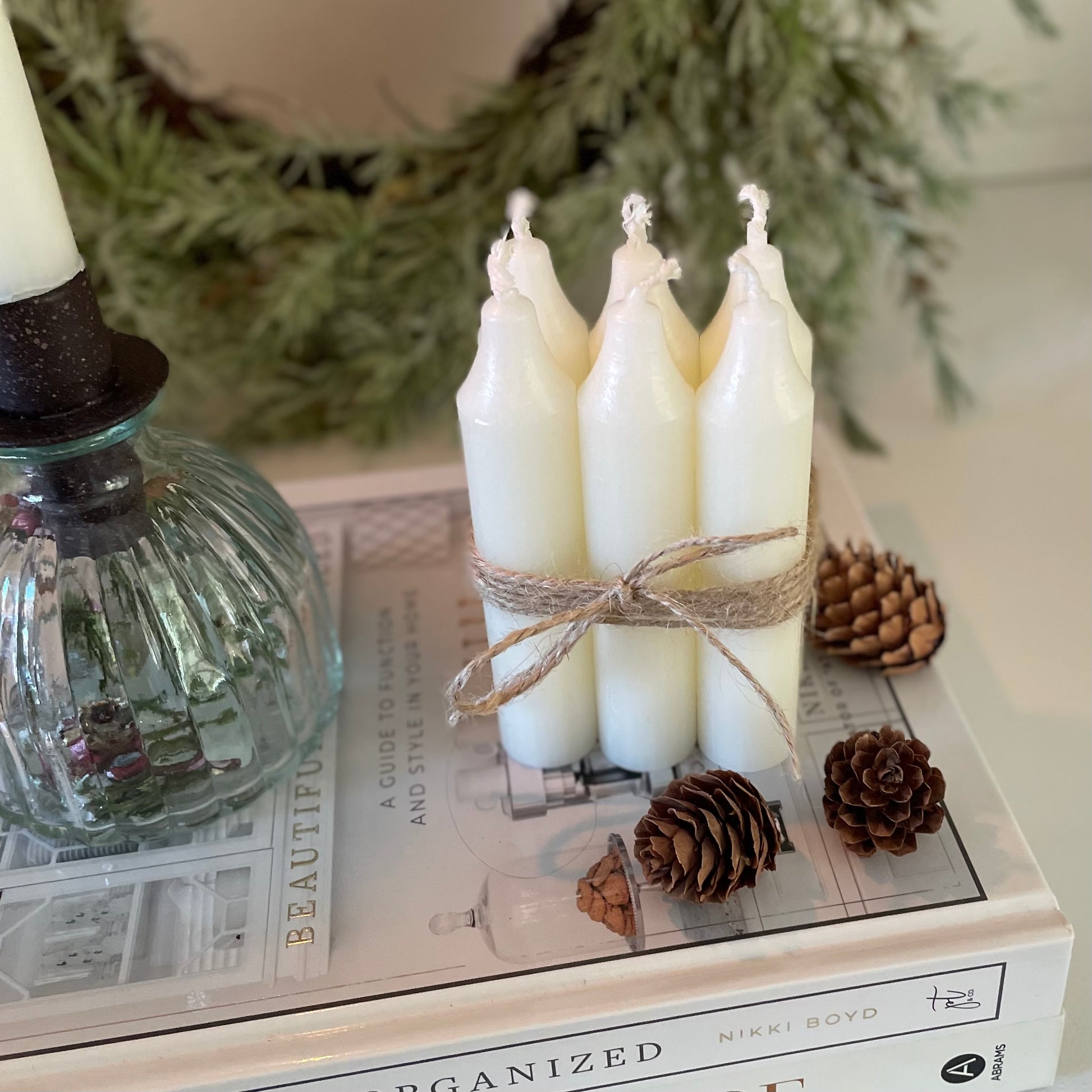 Bundle of six short Danish style dinner candles tied with jute cord, styled on a book with pine cones and candle holder.