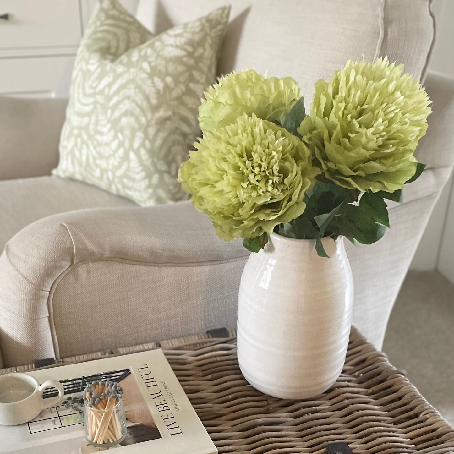 Single short peony stems styled in a vase on a wicker trunk with a book and cosy cream sofa.