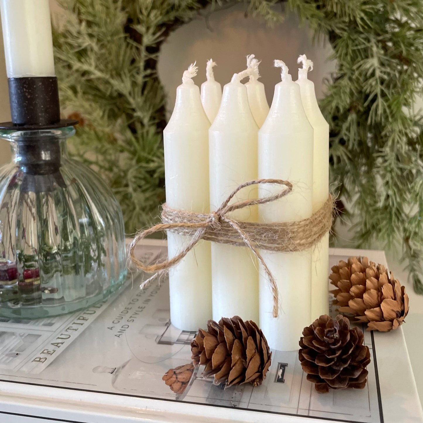 Bundle of six short Danish style dinner candles tied with jute cord, styled on a book with a wreath in the background.
