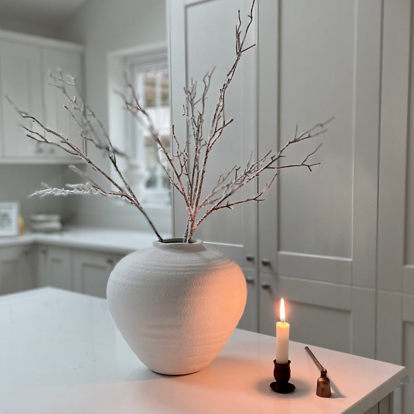Winter faux branch styled in a white matte ceramic vase next to a candle on a white  kitchen island.
