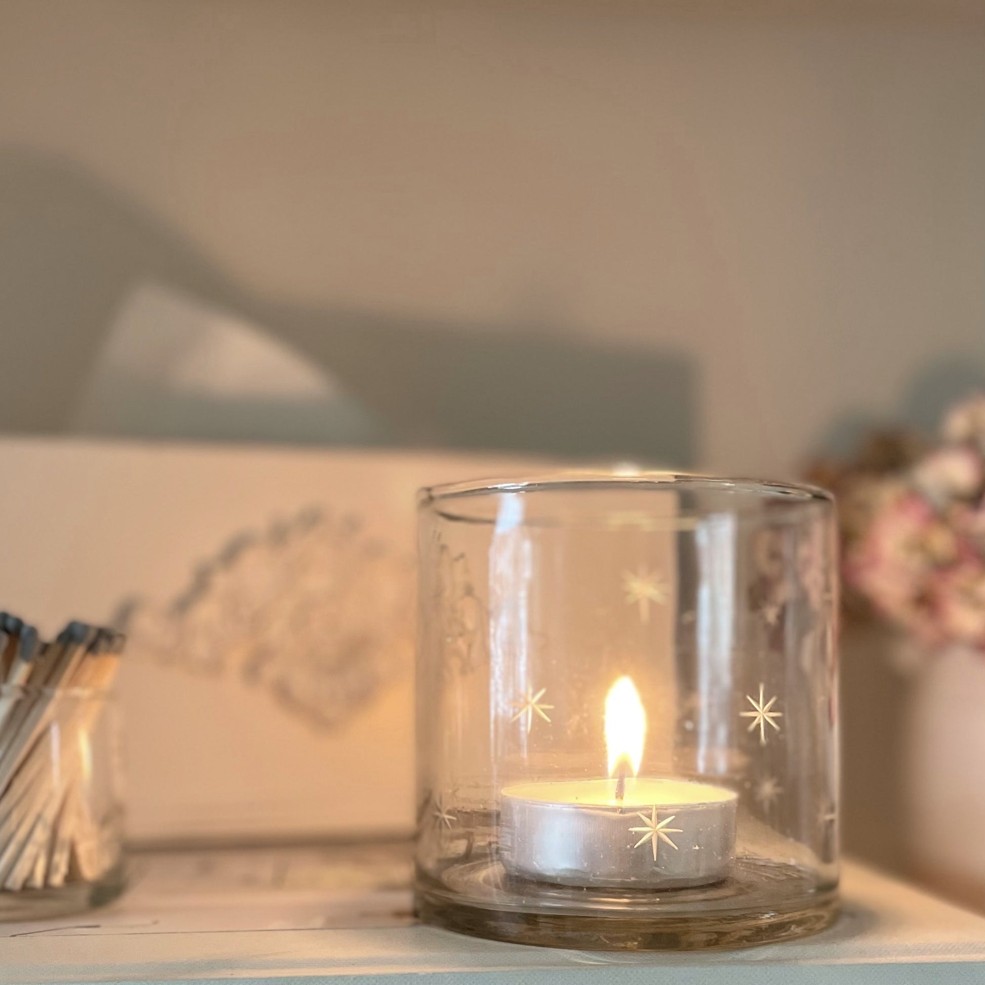 Glass hurricane candle holder with delicate etched star design, styled with a glowing candle on a book and shelf.
