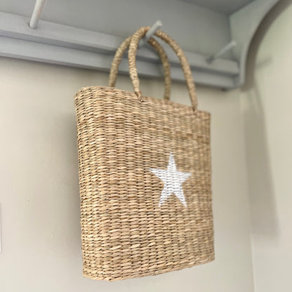 Hand-painted white star seagrass tote bag with natural woven handles, styled on a peg shelf.