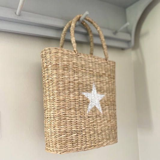 Hand-painted white star seagrass tote bag with natural woven handles, styled on a peg shelf.