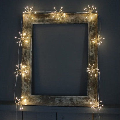 Warm white starburst string lights draped along a rustic picture frame, creating a soft festive glow indoors.