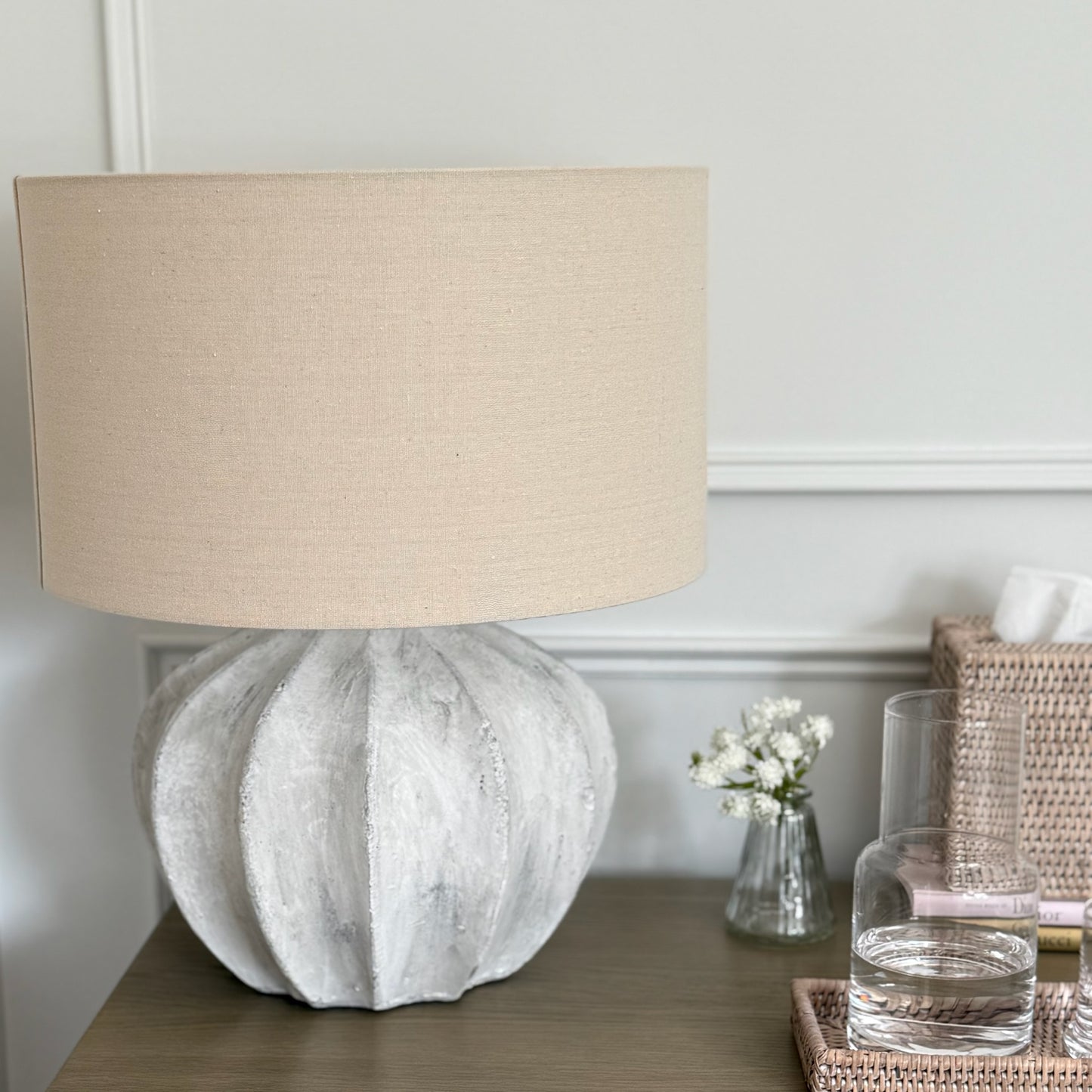 Table lamp with beige shade on a wooden bedside cabinet, with a vase and glass in the background.