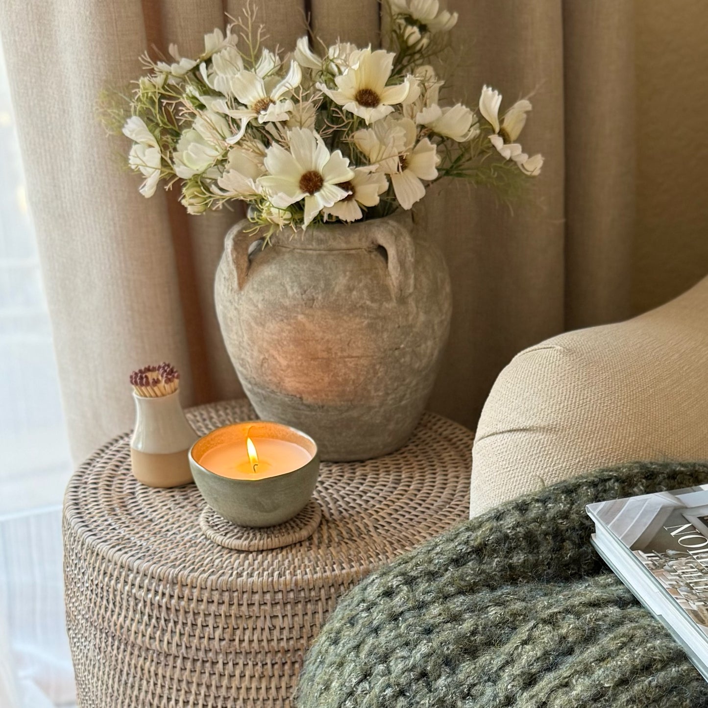 Vase with flowers on a woven side table next to a lit candle, with a couch and magazine in the background.