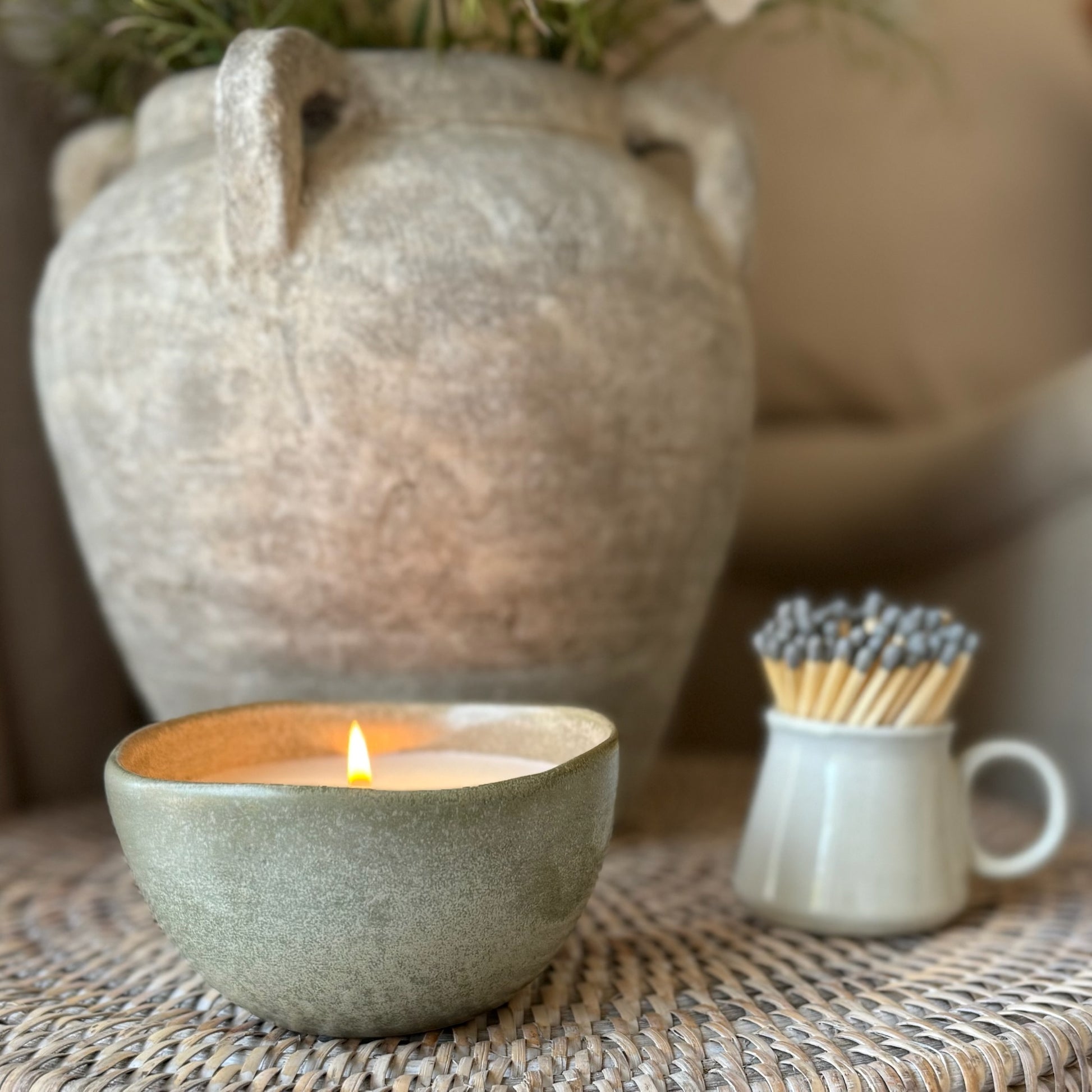 Ceramic bowl with a lit candle, matches, and a textured vase on a woven surface.