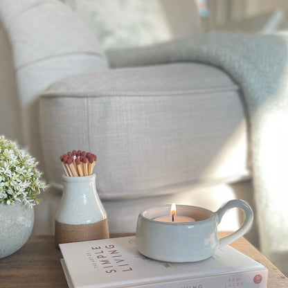 White ceramic tea light holder with handle, styled on a wooden stool and book with a hebe plant for a cosy home look.