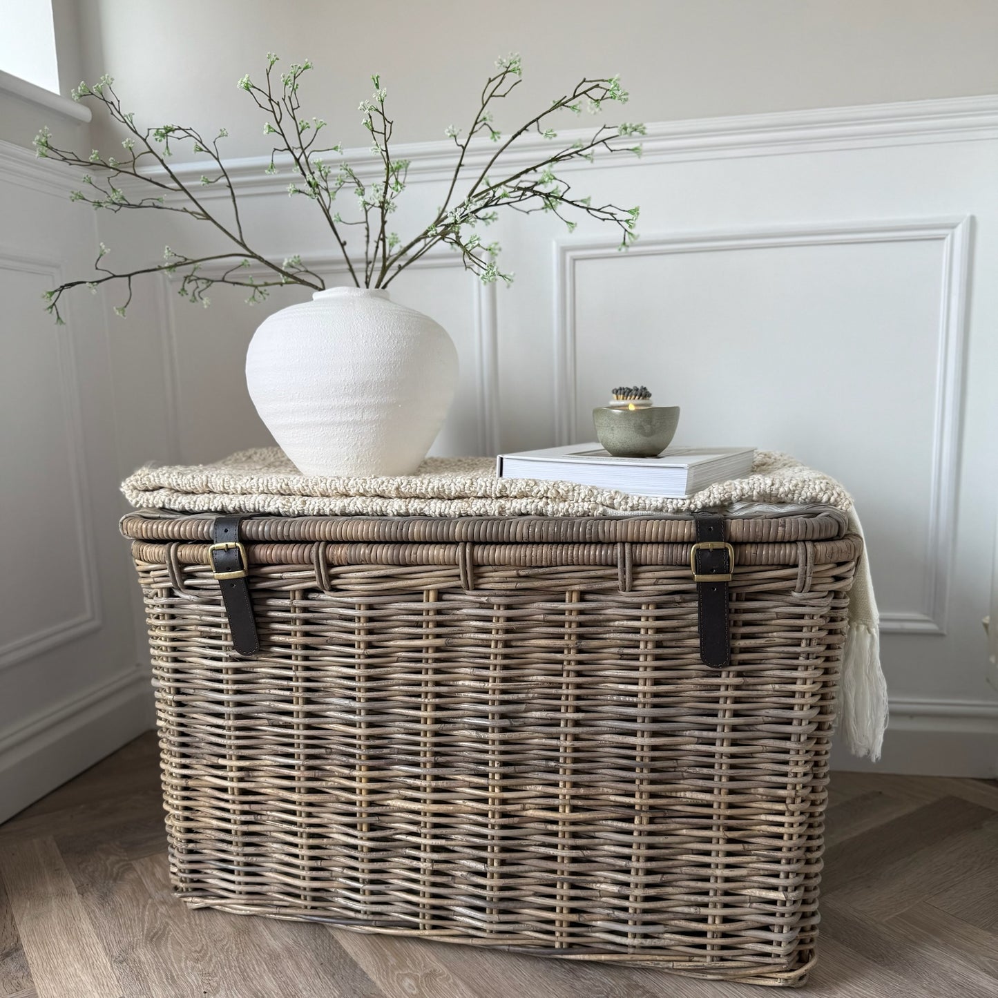 Wicker storage trunk with a white vase and books styled in a hallway on a wooden floor.