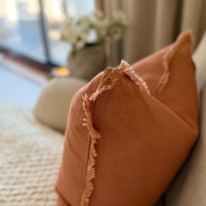 Close-up of a terracotta coloured cushion with frayed edges on a blurred indoor background.