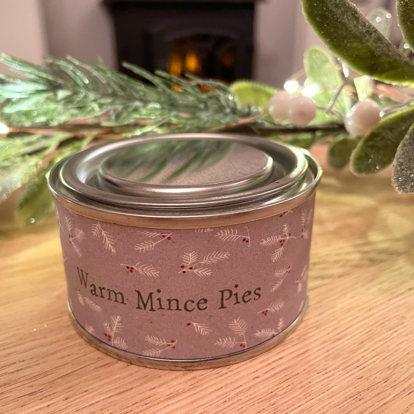 Warm mince pies scented christmas tin candle, styled on a wooden coffee table with a green garland.