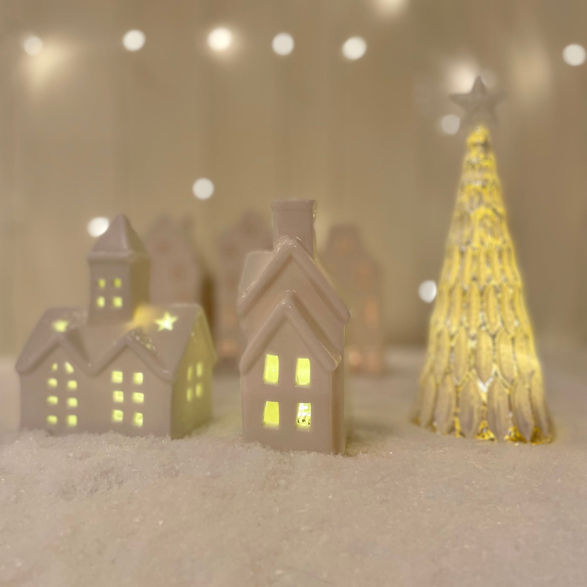 Small white glazed ceramic LED house ornament with warm light, styled on a snowy scene with other ceramic christmas decor.