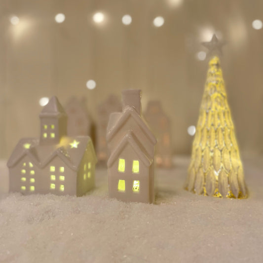 Small white glazed ceramic LED house ornament with warm light, styled on a snowy scene with other ceramic christmas decor.