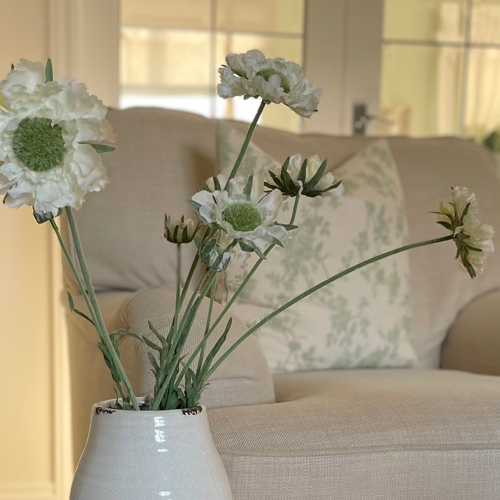 Faux Scabiosa Stem with off white flowers and soft green centres, styled in a glazed vase. Placed on a stool in a cosy living room.