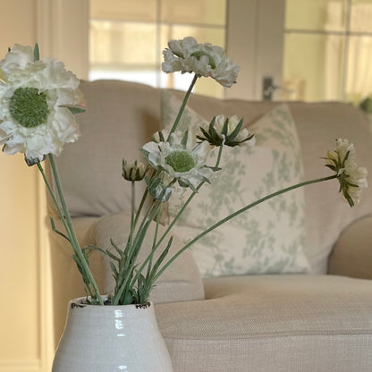 Faux Scabiosa Stem with off white flowers and soft green centres, styled in a glazed vase. Placed on a stool in a cosy living room.