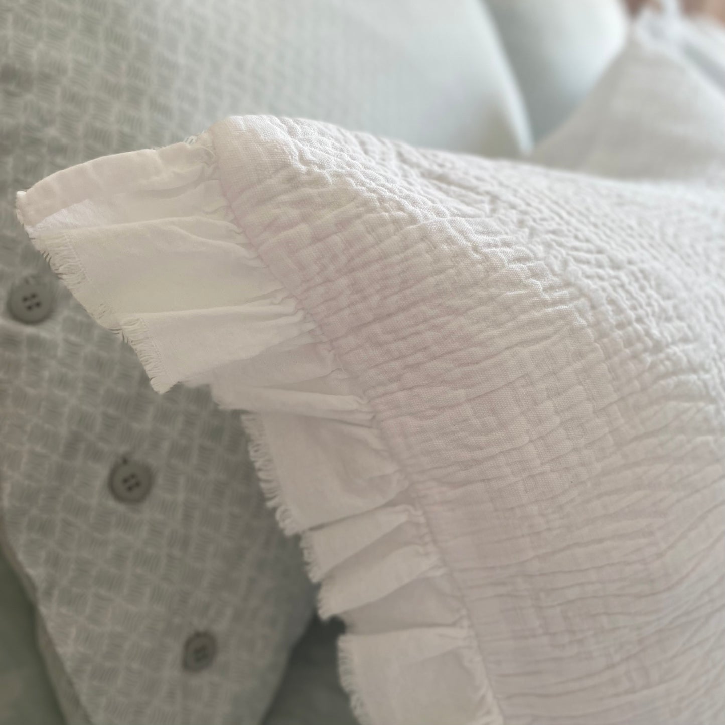 Close up of white quilted cushion with frilled edges and embroidered textured design. Styled on a bed.