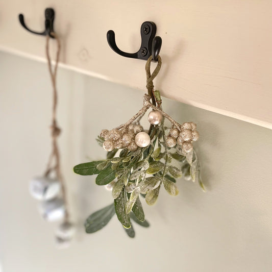 Glittered mistletoe sprig with white berries hanging from a rustic hook against a festive background.