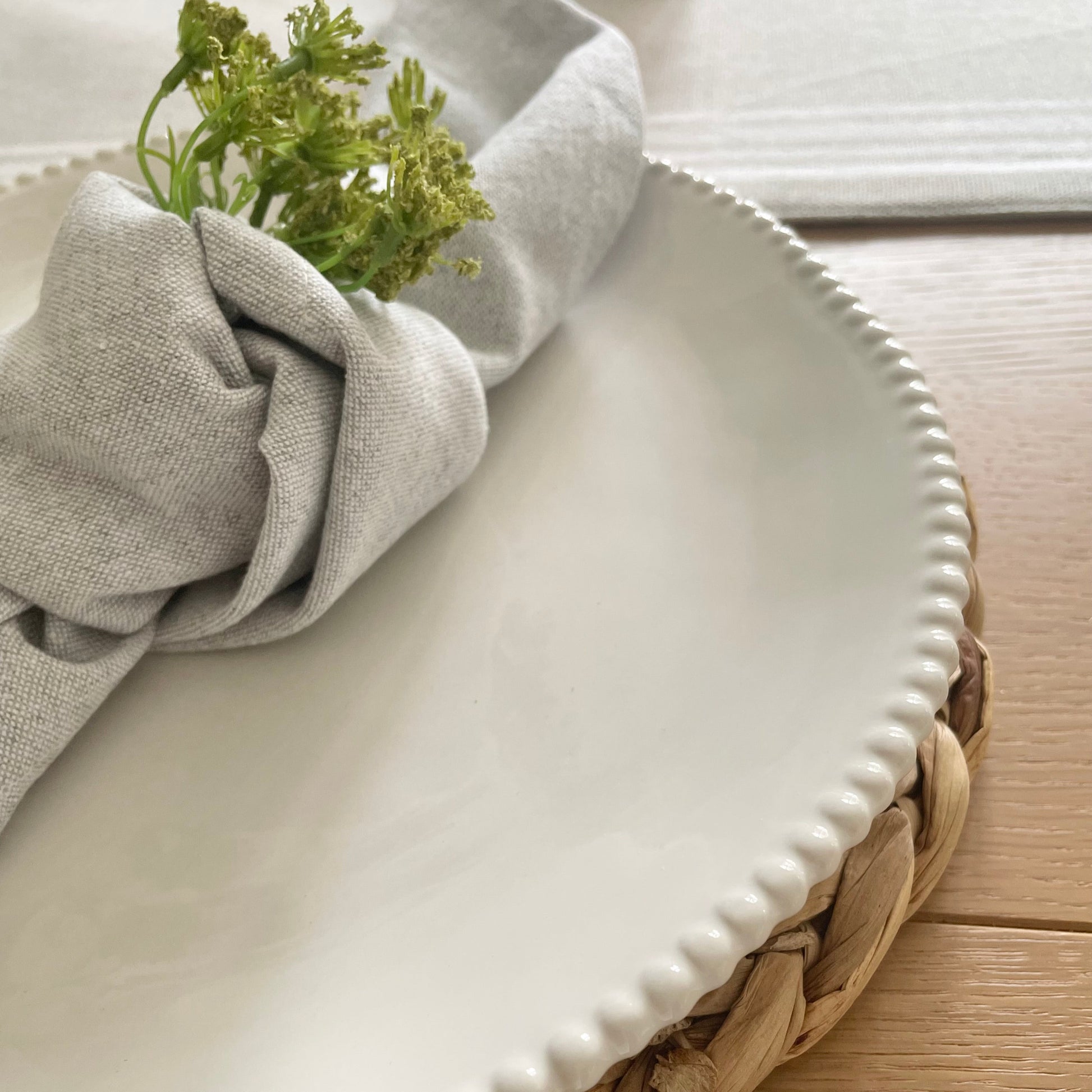 White porcelain dinner plate with organic shape and beaded detailing around the rim, styled on a dining table with grey napkin and seagrass placemat.