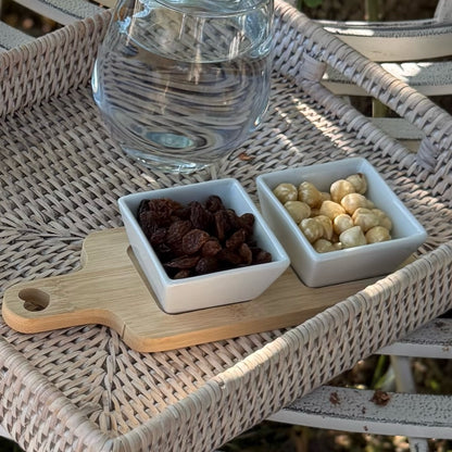 Two small bowls with raisins and nuts on a wooden board with a glass of water on a wicker table.