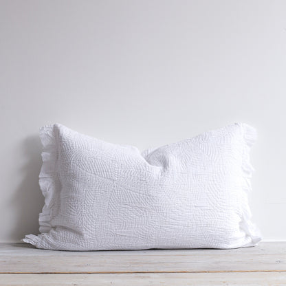 White quilted cushion with frilled edges and embroidered textured design. Styled on a wooden bench.