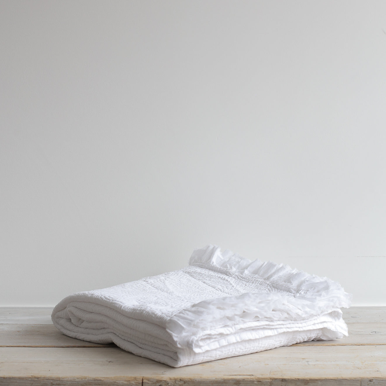 White quilted bedspread with embroidered texture and frilled edges. Styled on wooden bench.