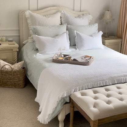 White quilted cushion with frilled edges and embroidered textured design. Styled on a bed with co ordinated throw. In a clam bedroom setting.