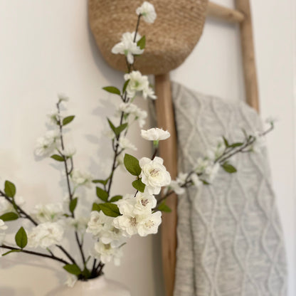 Decorative arrangement with white faux blossom flowers, a woven basket and a cable knit blanket against a white wall.