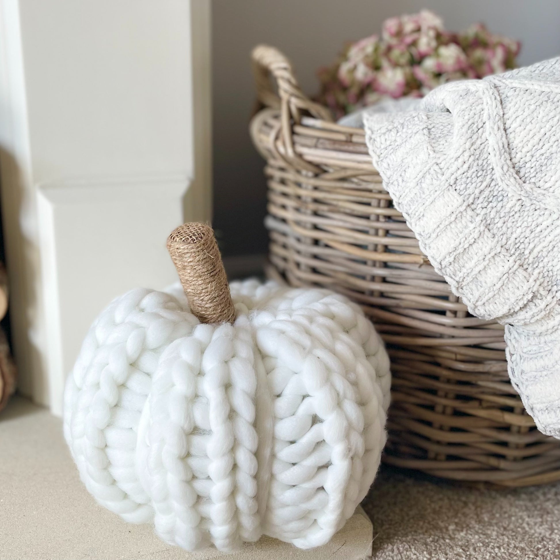 White chunky knitted pumpkin with hessian stalk styled next to a rattan basket with cable knit throw.