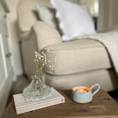 White linen cushion with ruffled edge and mother of pearl button detail styled on a chair with a throw and stool with candle and book.