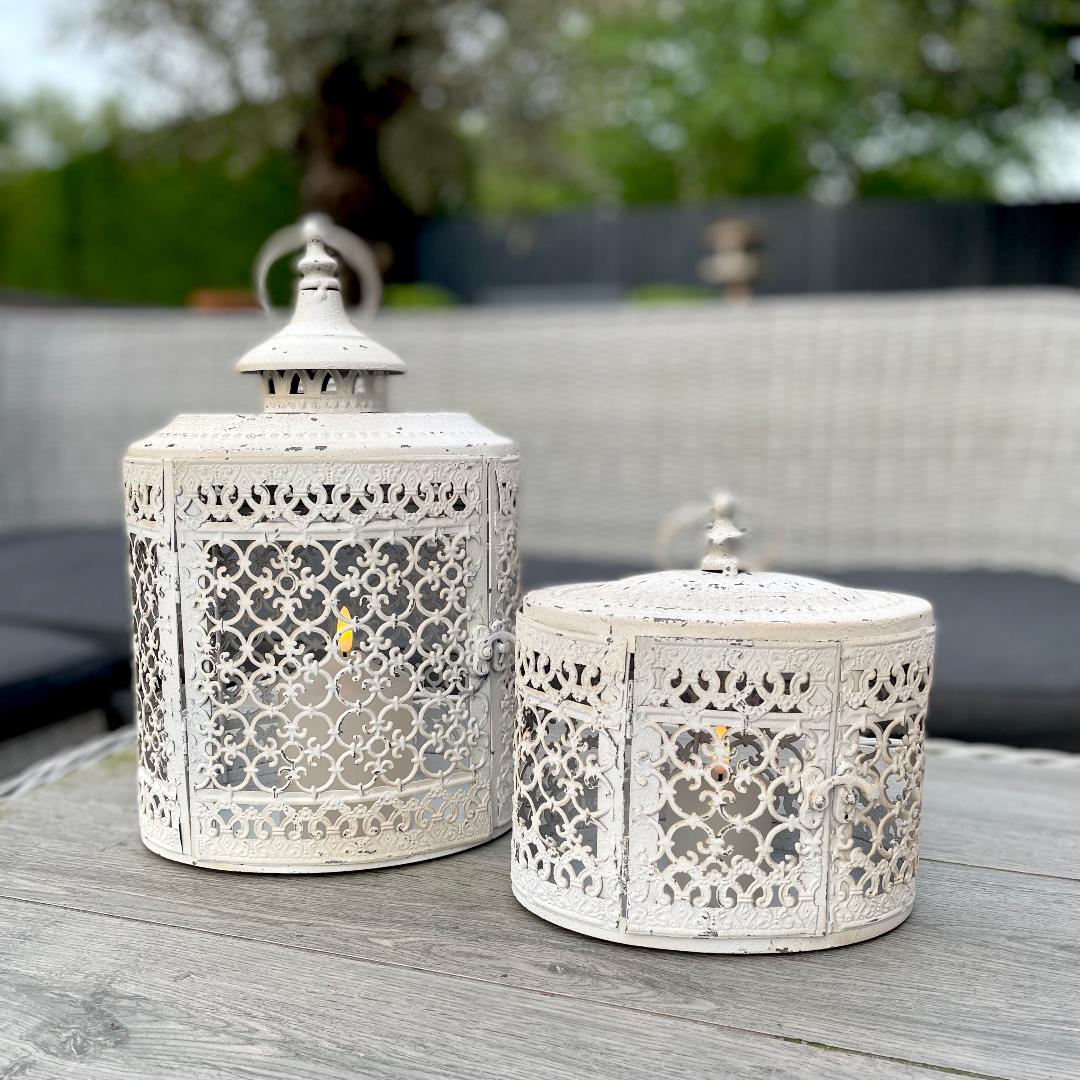 Antique white oval metal lantern with vintage finish casting soft candlelight in a cosy garden setting.