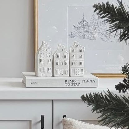 Decorative set of three white houses, styled on a book on a console table with a framed picture in the background. Christmas tree and chair in background.