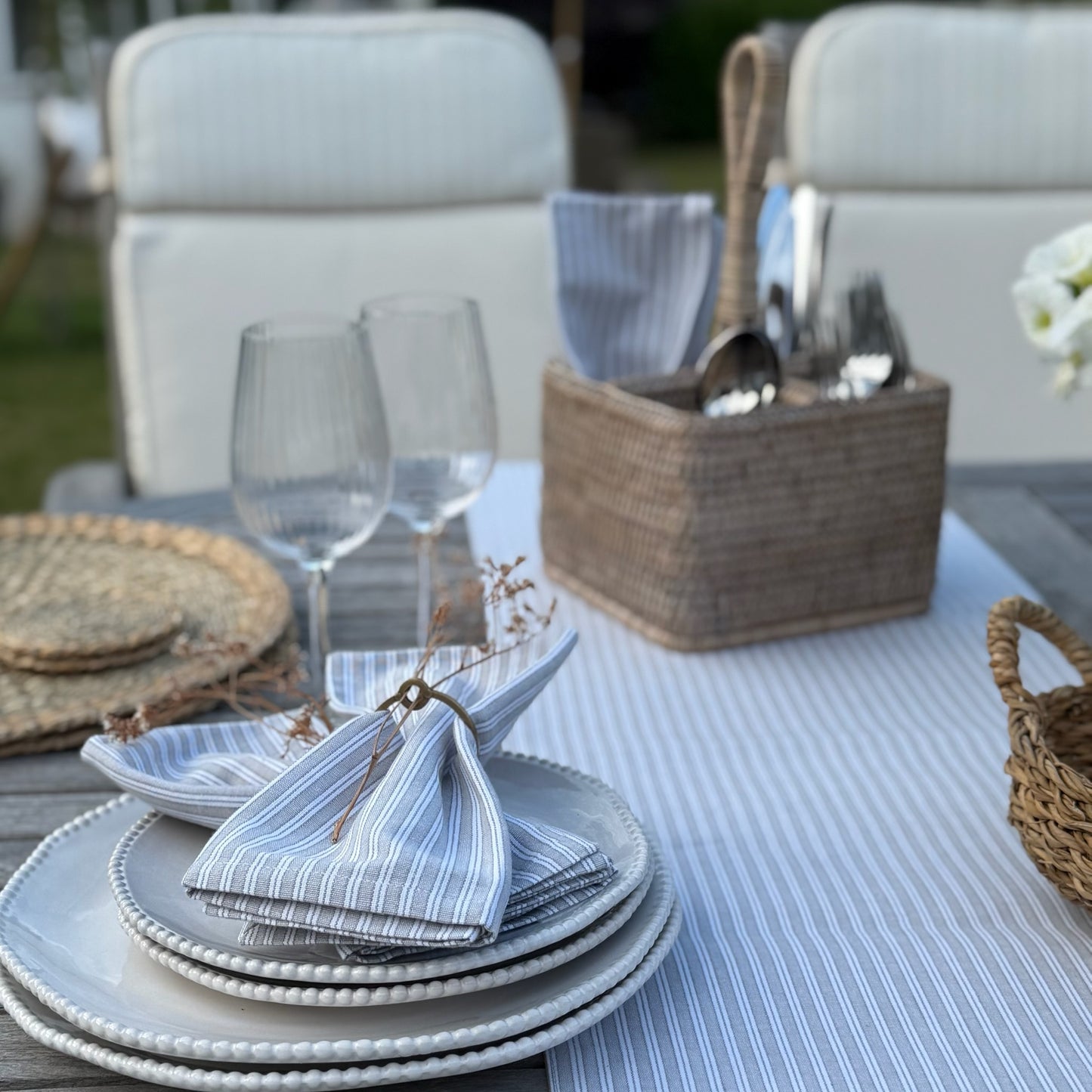 Ethically sourced natural rattan caddy with carry handle. Dining set up with plates, glasses and cutlery on a striped tablecloth.