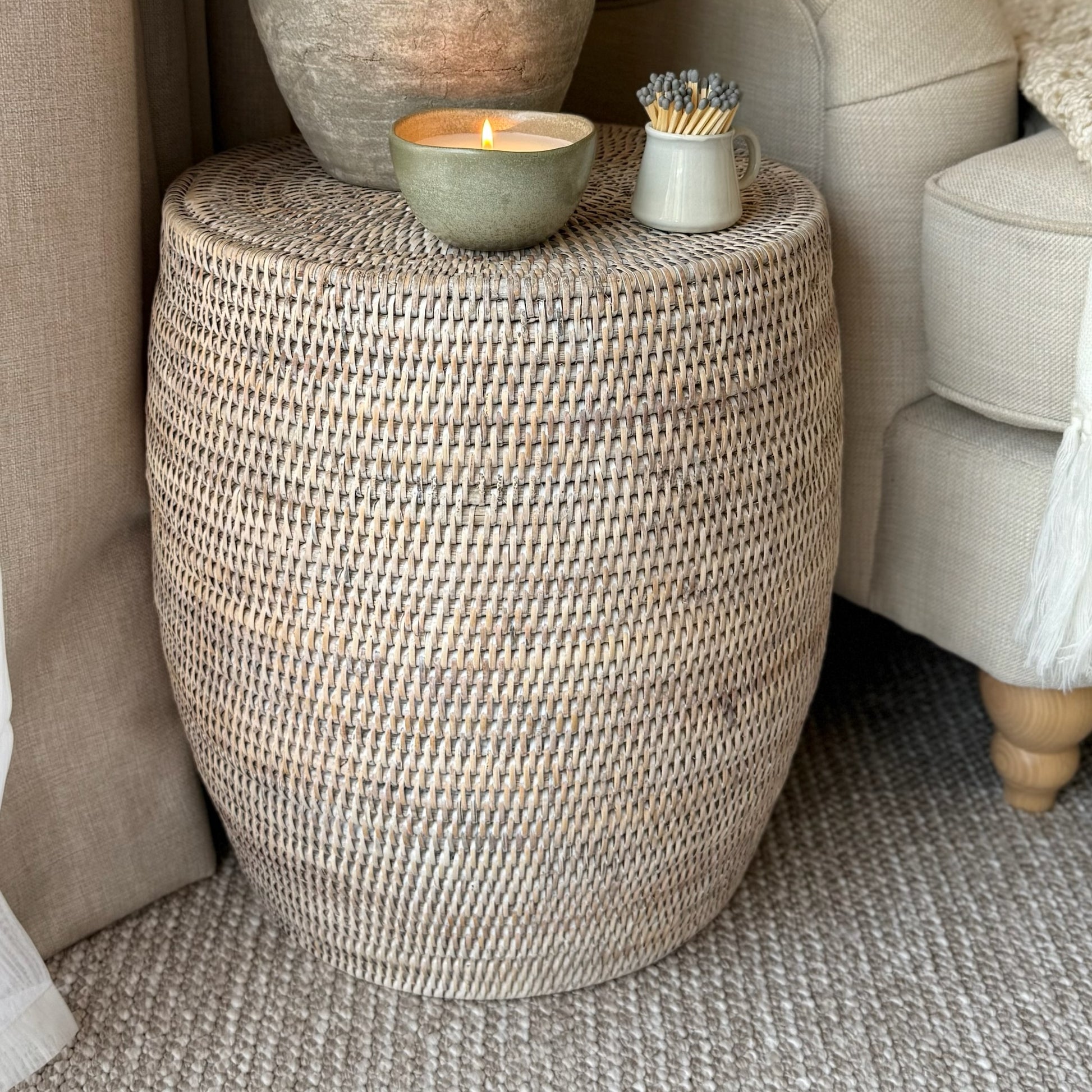 Handwoven white-washed rattan drum stool, styled as a side table with books and a plant in a calm, neutral interior.