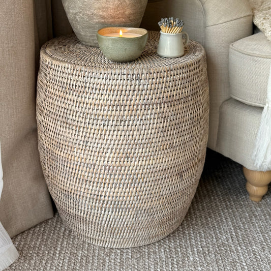 Handwoven white-washed rattan drum stool, styled as a side table with books and a plant in a calm, neutral interior.