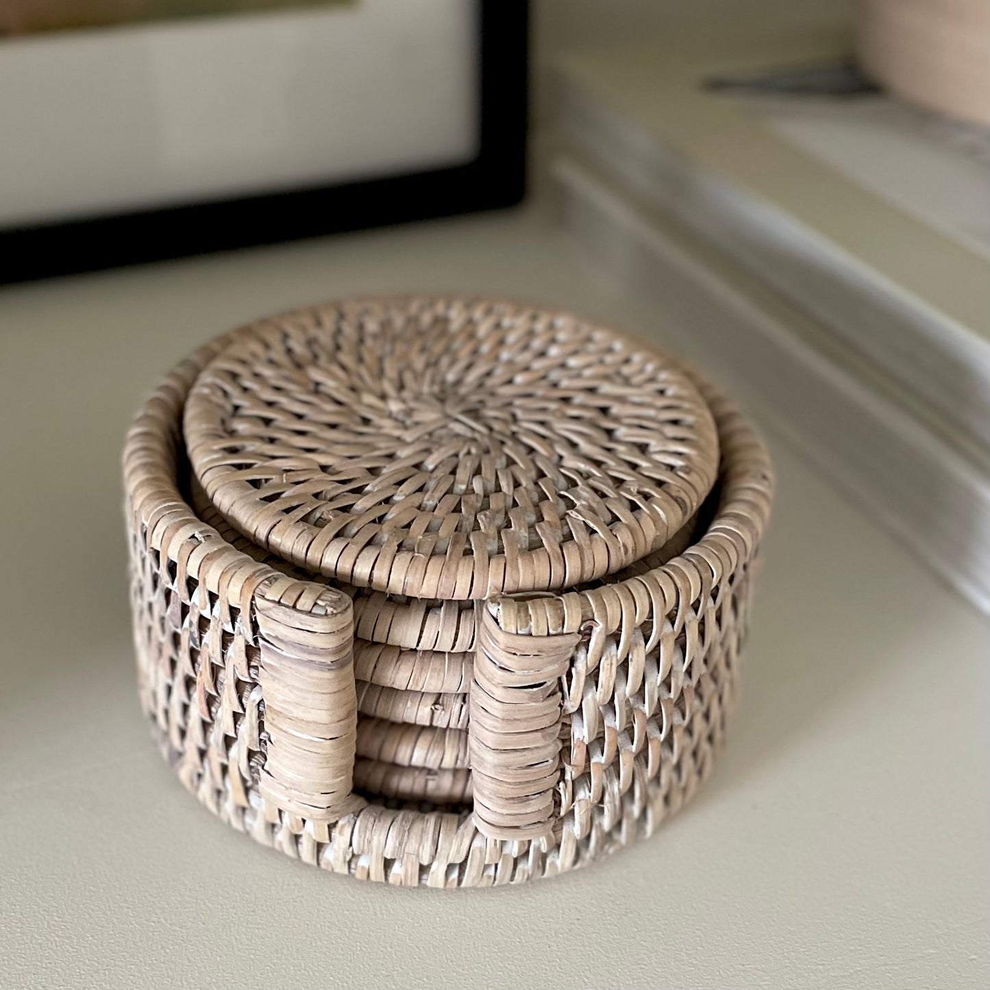 Set of 6 handcrafted whitewashed rattan coasters with holder. Styled on a shelf with other decorative items.
