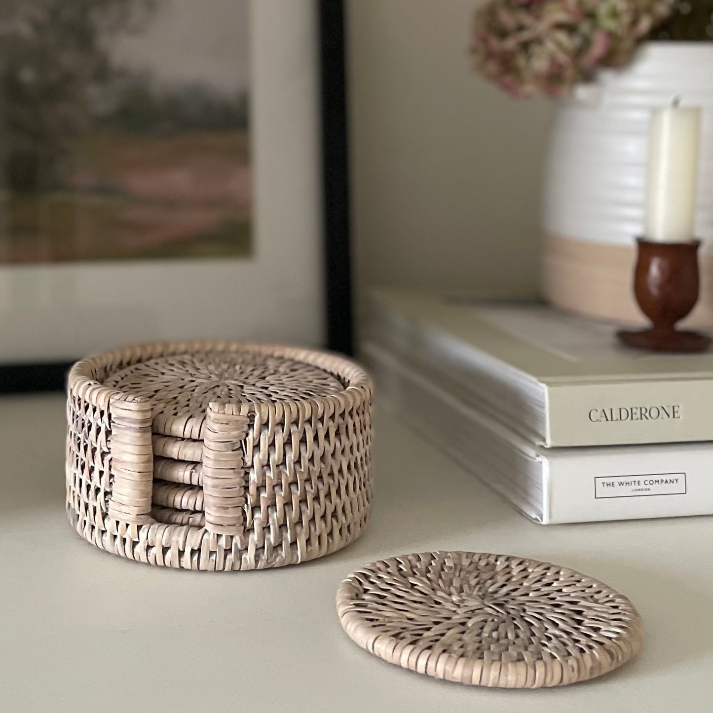 Sustainable and ethically sourced rattan coaster set. Styled on a shelf with books, candle and pot of hydrangea dried flowers.