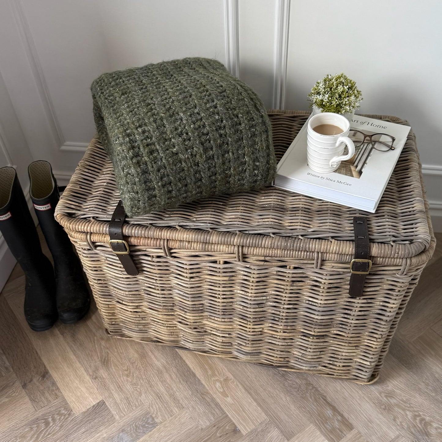 Wicker storage trunk with a green blanket, coffee cup and book on a wooden hall floor.