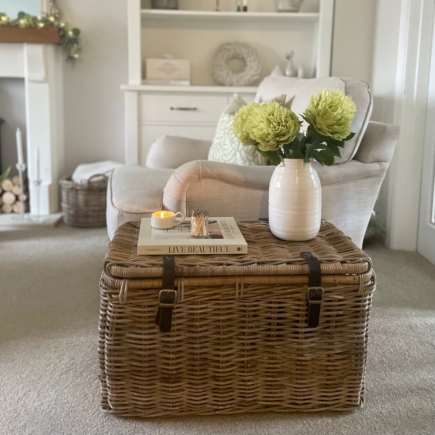 Wicker storage trunk with brown leather straps styled in a cosy living room with a vase of flowers.