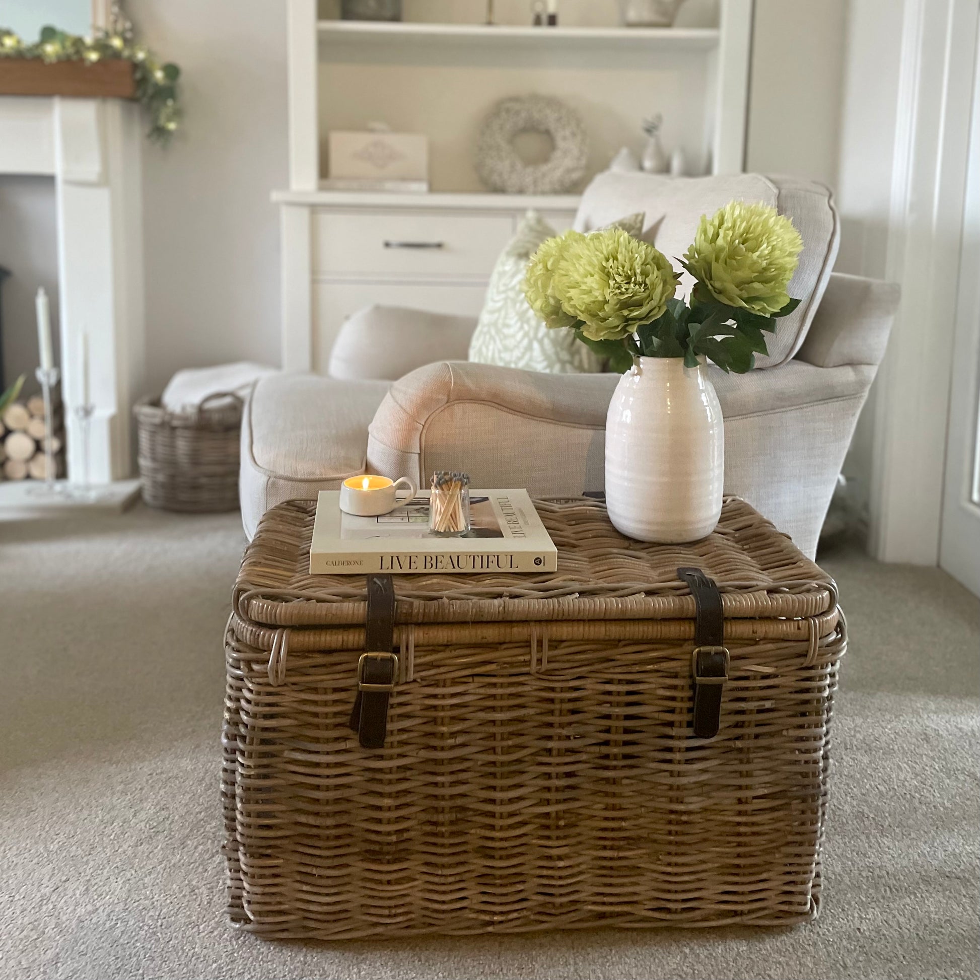 Wicker storage trunk with brown leather straps styled in a cosy living room with a vase of flowers.